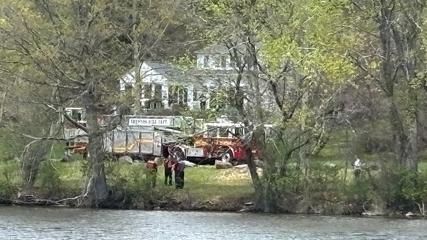 FireTruckMissingStudent.jpg A fire truck is parked on the grass next to a lake, a white house is in the background.
