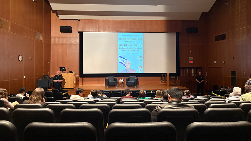 Lots of seats are empty in a wood-paneled classroom with a screen and podium up front.
