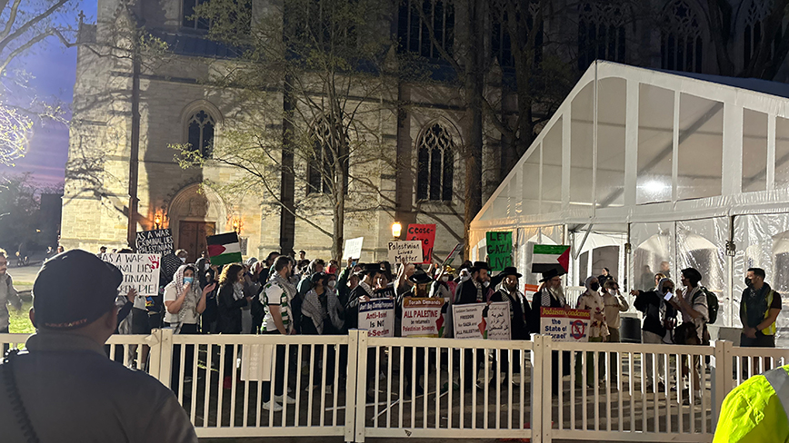 People hold signs behind a white fence with the University Chapel in the background.