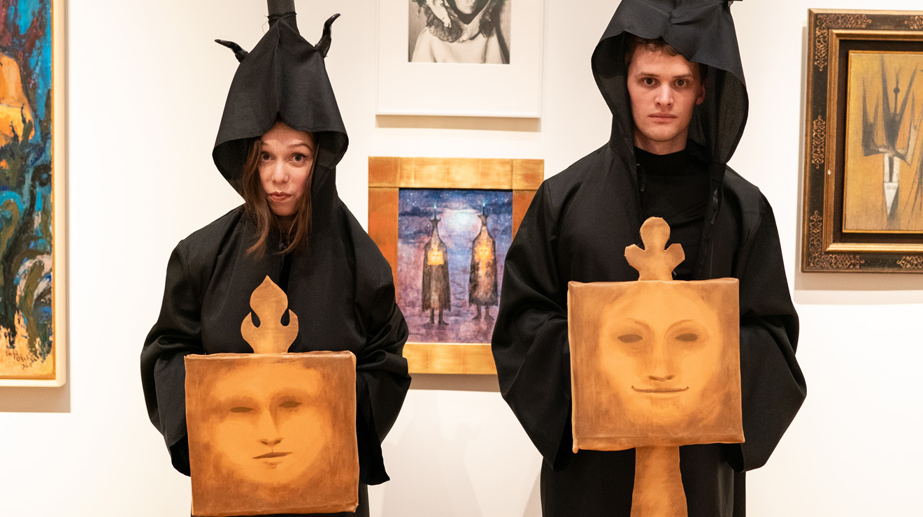 PAUM OPENING_Dark Hoods.jpg Two people wearing dark robes and hoods hold paintings of faces in an art museum..
