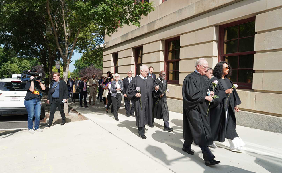 Judges in black robes walk along a sidewalk next to a building while a two-person TV crew films them.