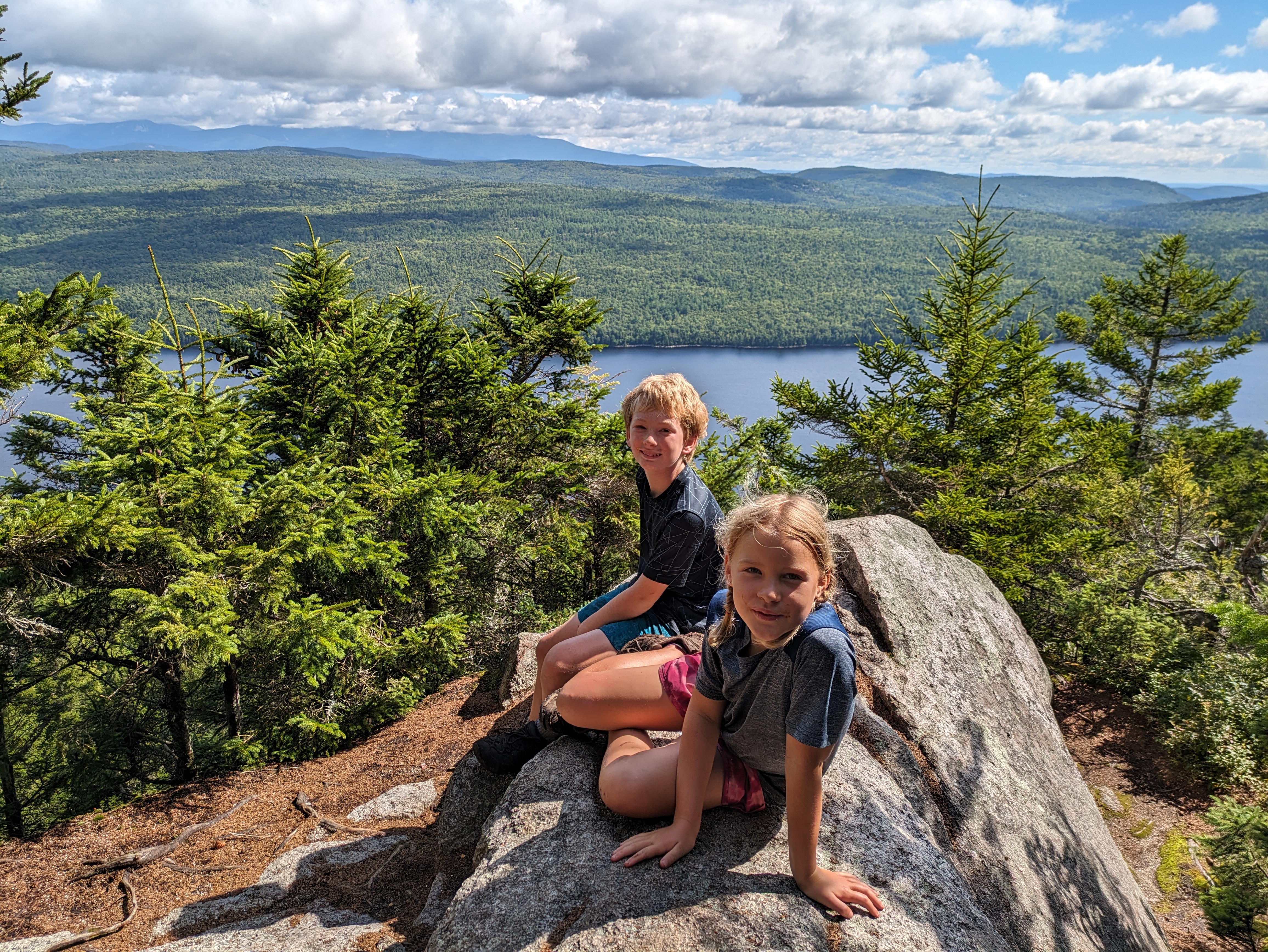 PXL_20230814_140845636.jpg Two children sit on a rock high above a body of water, either a lake or a river.