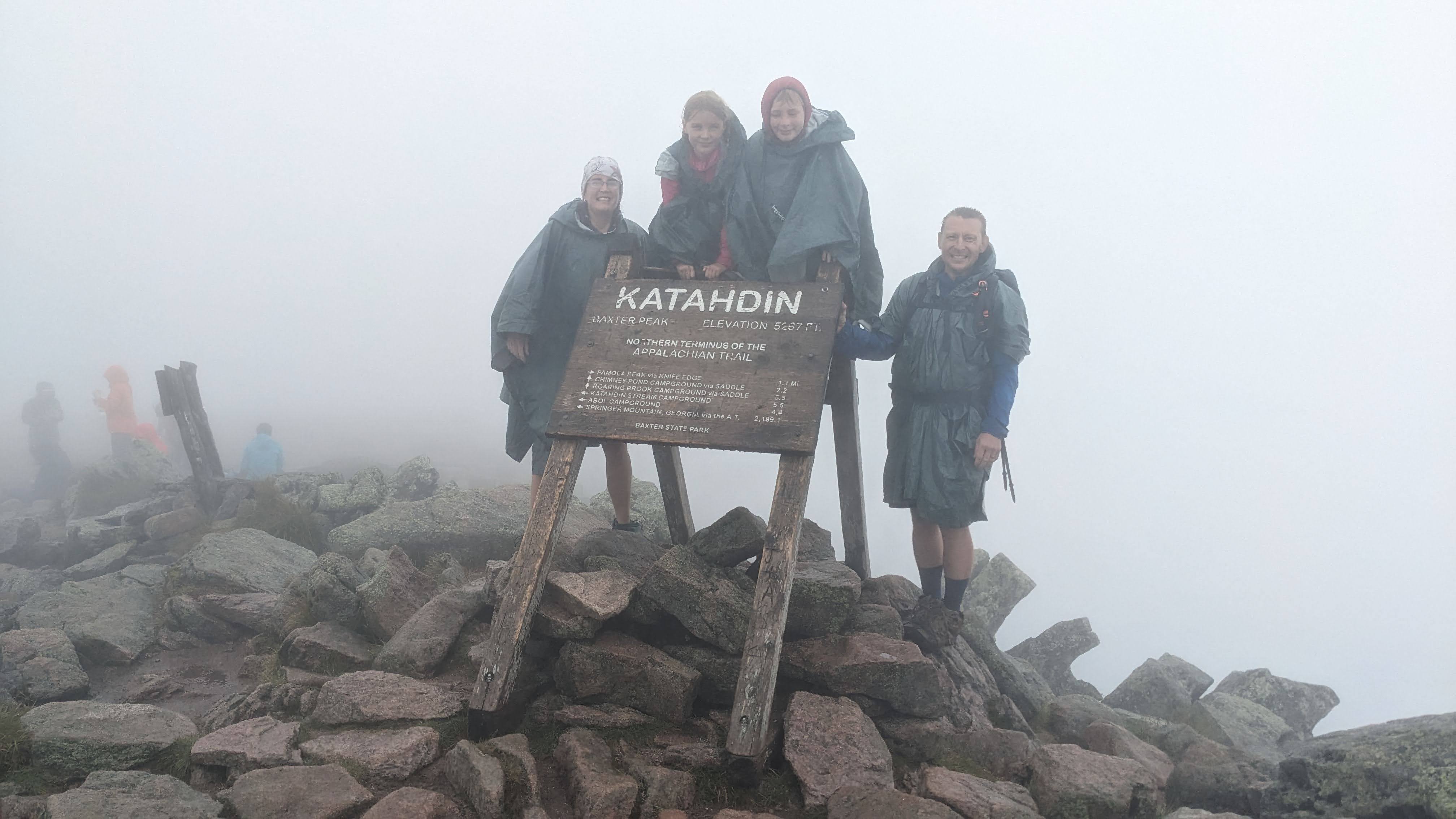 PXL_20230817_172542479.jpg Four people in rain jackets cluster in thick fog around a wooden sign reading “Katahdin,” perched on a pile of rocks.