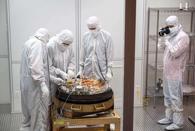 STAR MAN_Cracking the case.jpg Four people in white hazmat suits stand around the sample from asteroid Bennu as it's unpacked