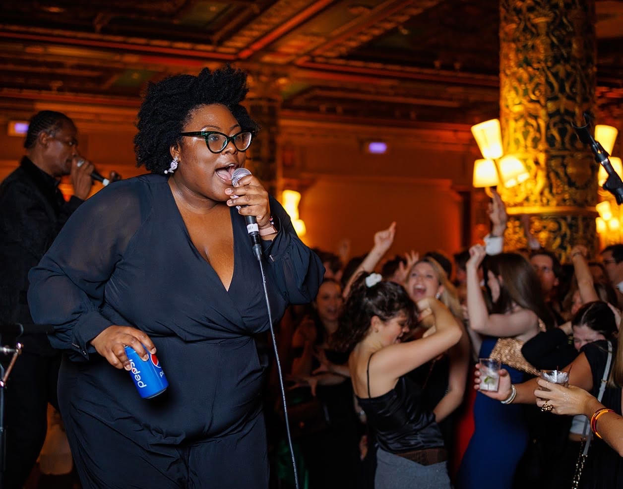 A woman sings into a microphone; people dance in the background.