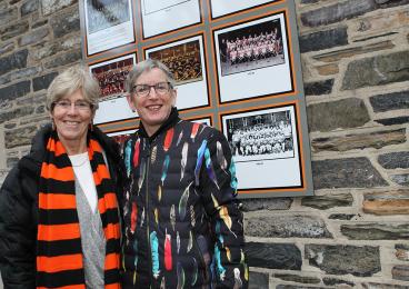 Carol Brown ’75, left, and Elizabeth English ’75 stand in front of a wall of hockey team photos on a stone wall at Baker Rink.