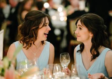 Kate and Tori wearing matching blue dresses and holding wine glasses at a table during a wedding reception.