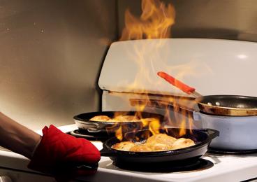 This is a photo of a student's hand wearing an oven mitt shaking a flaming skillet over a dorm-size stove.