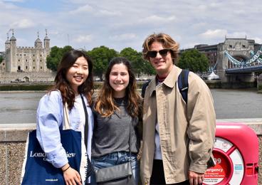 Three students stand on a bridge with the Tower of London and Tower Bridge in the background.