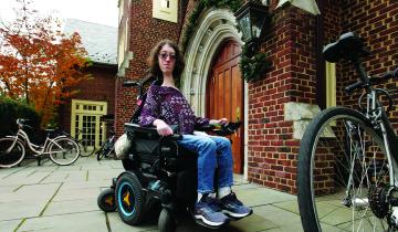 This is a photo of Naomi Hess ’22 in her motorized wheelchair in front of a Princeton campus building. Bicycles are in the foreground and background.