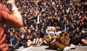 The Class of 2017 gathers in their black jackets behind the Tiger mascot, who's sitting on the ground and clapping his paws.