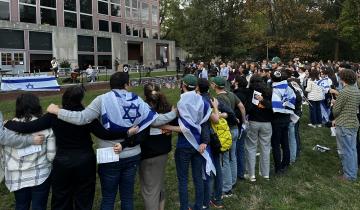 People stand with their arms around each other's shoulders outside a campus building, listening to two students play music. A few have blue-and-white Israel flags wrapped around their shoulders.