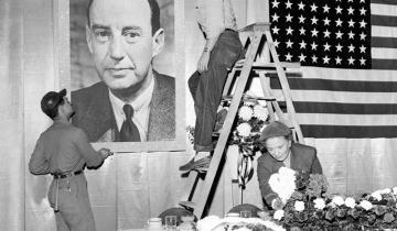 In this black-and-white photo, two men hang a poster of Adlai Stevenson on a wall next to an American flag, while a woman arranges flowers on a long table. Preparations are made for a campaign stop for Adlai Stevenson 1922 in 1952, before he lost to Dwight Eisenhower, 442 electoral votes to 89.