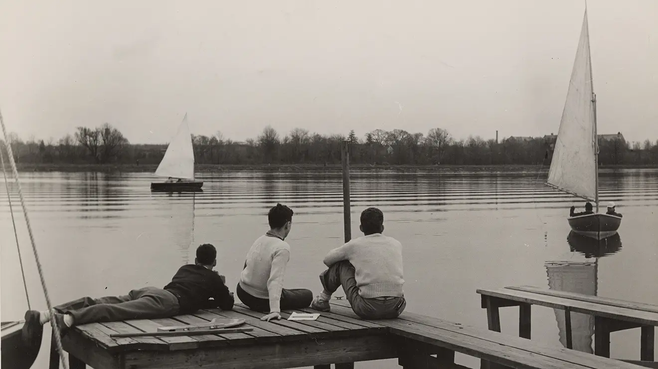 Three students sit at a dock and look at sailboats on Lake Carnegie in this sepia-toned photo.