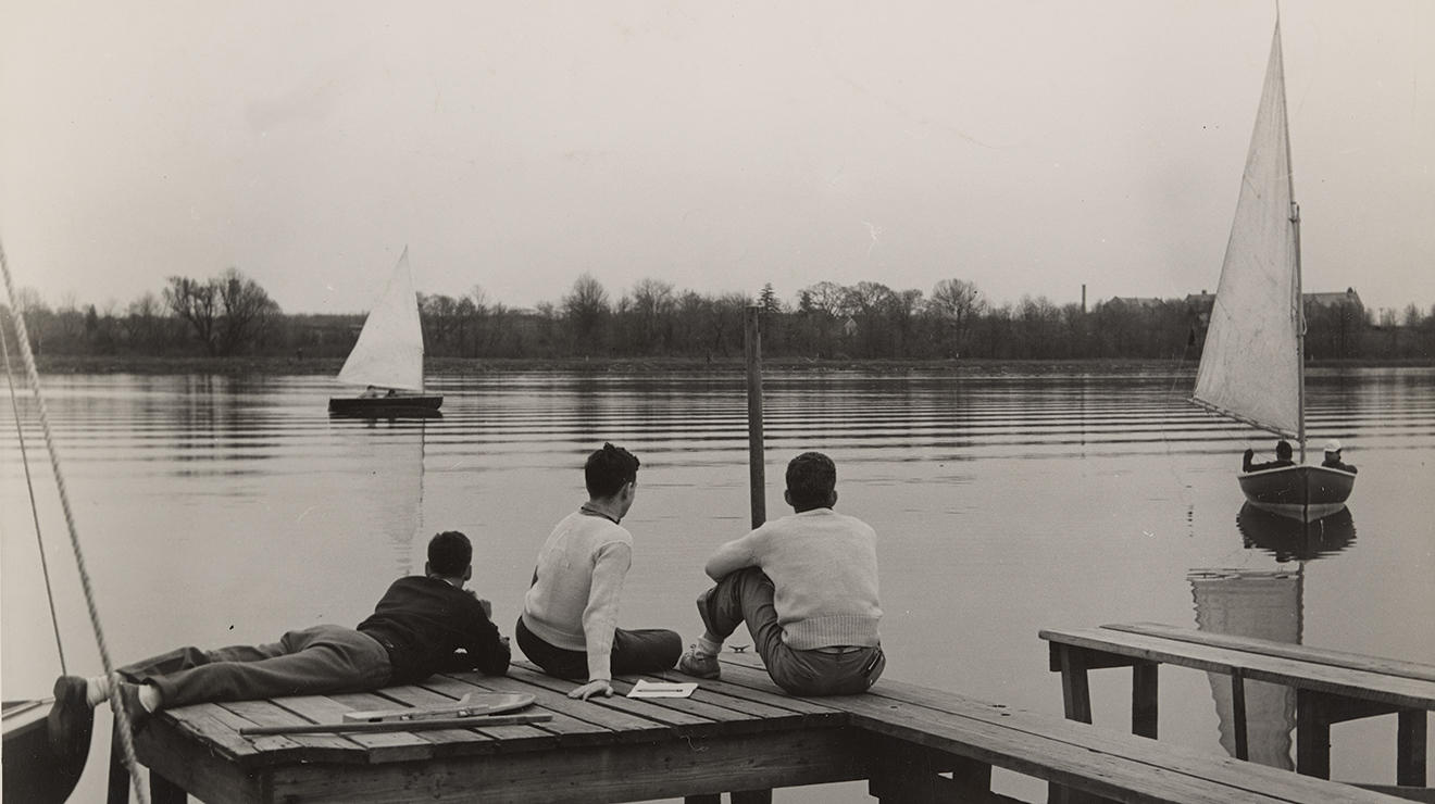 Three students sit at a dock and look at sailboats on Lake Carnegie in this sepia-toned photo.