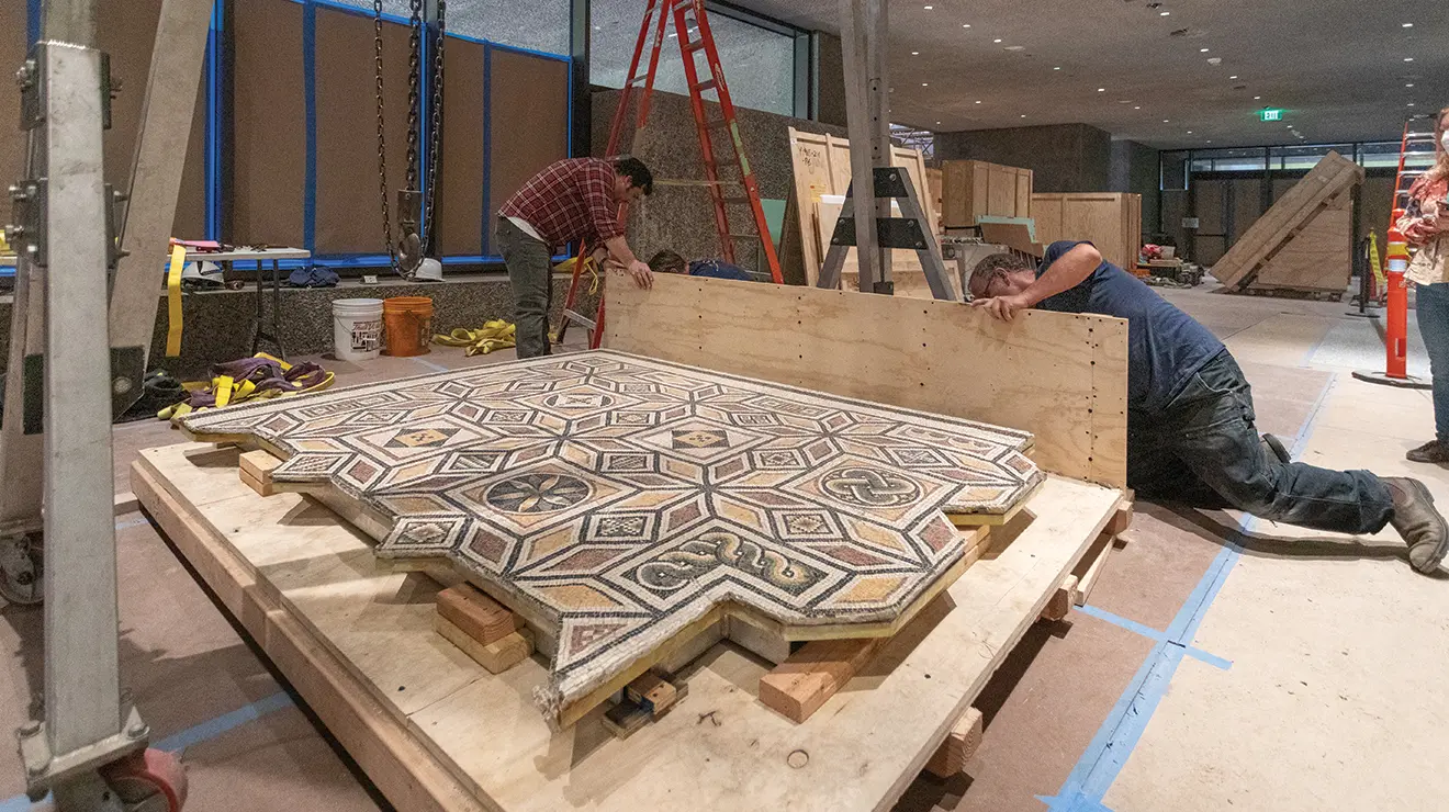 Two men work on a wood platform holding a Roman floor mosaic.