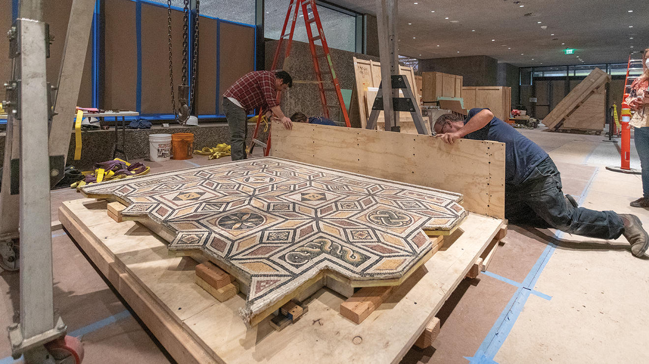 Two men work on a wood platform holding a Roman floor mosaic.
