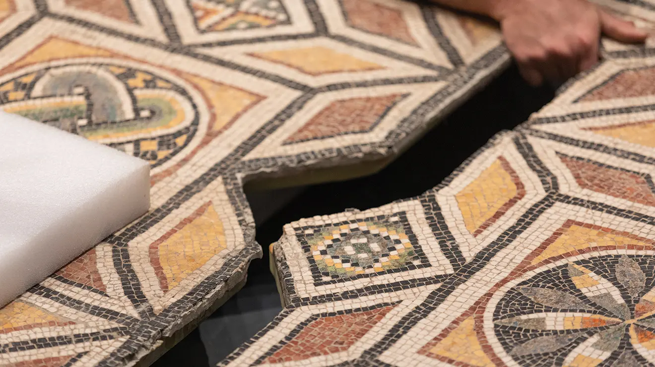 Close up, a hand pulls one side of a Roman floor mosaic into the other, like fitting puzzle pieces.