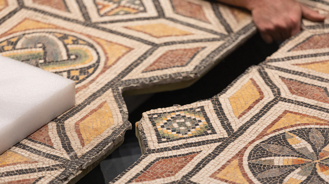 Close up, a hand pulls one side of a Roman floor mosaic into the other, like fitting puzzle pieces.