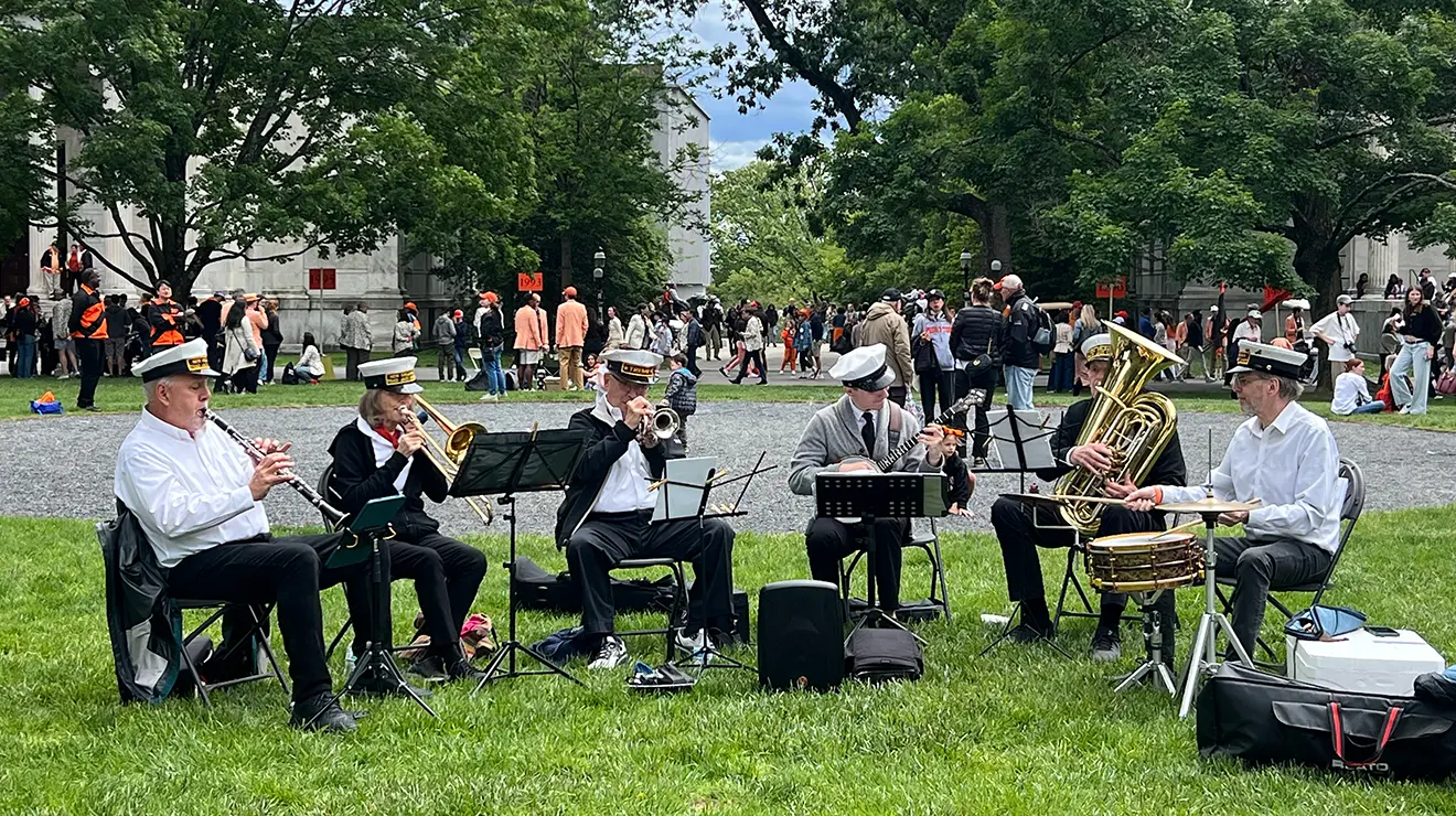 Six men wearing sailor hats play instruments, sitting in a semicircle outdoors.