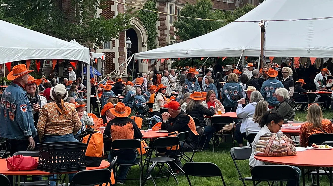 A crowd of people in orange cowboy hats hangs around tables under white tents.