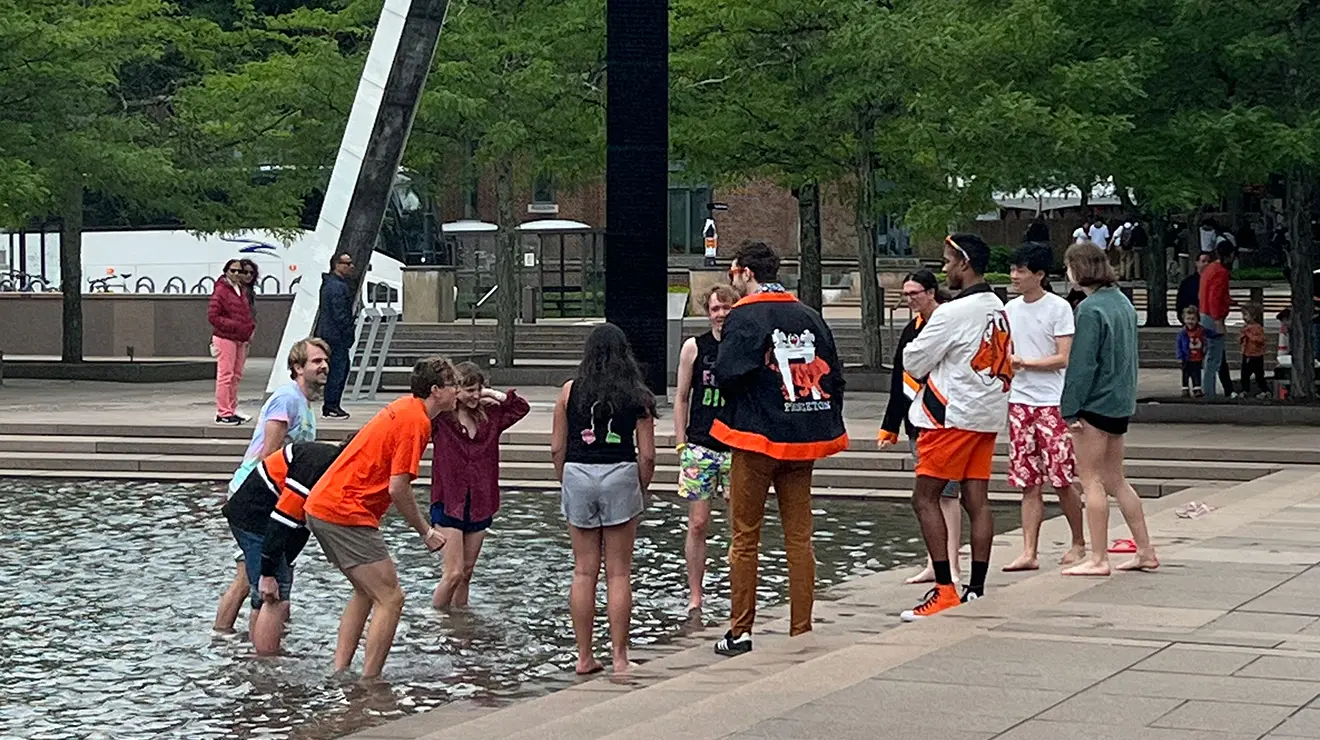 About a dozen young people form a circle at the edge of a fountain; half are in up to their ankles.