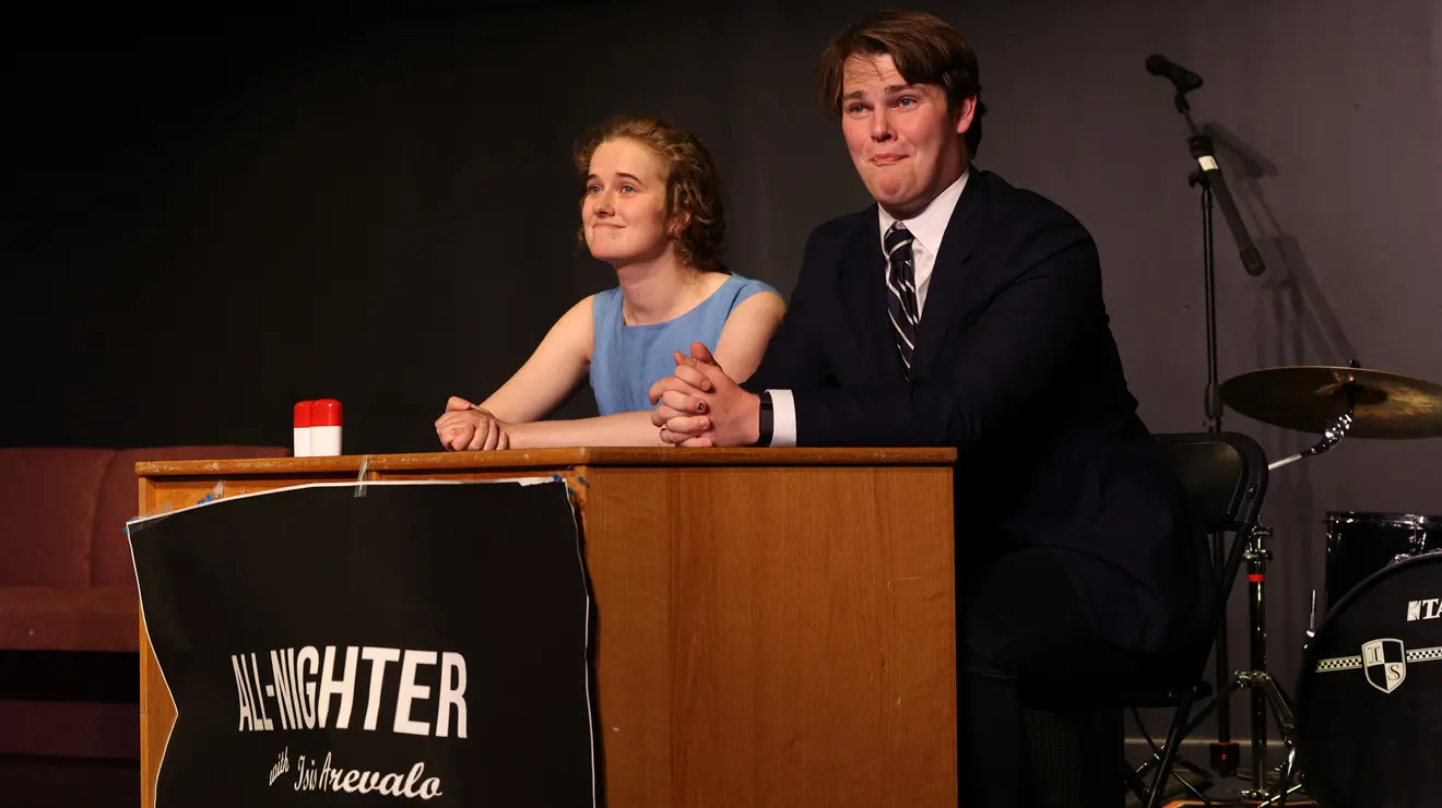 Two students sit at a dais with a drum set behind them. A sign in front says "All-Nighter."