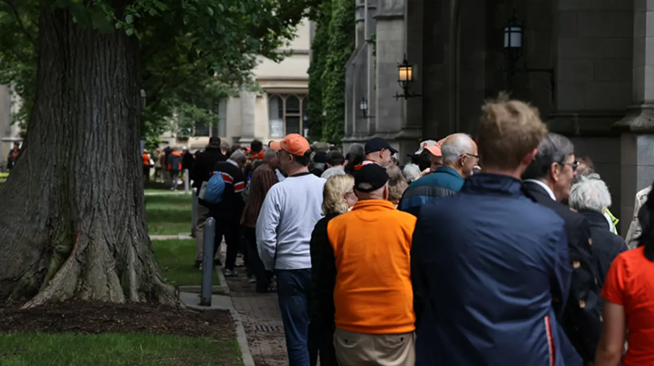 A large crowd of people waits in line outdoors, between a stone building and a tree, to see Gen. David Petraeus speak at Reunions.