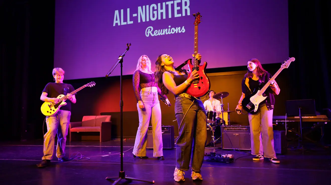 A female lead guitarist for a band holds her guitar in the air while performing in front of a screen that reads "All-Nighter: Reunions.".