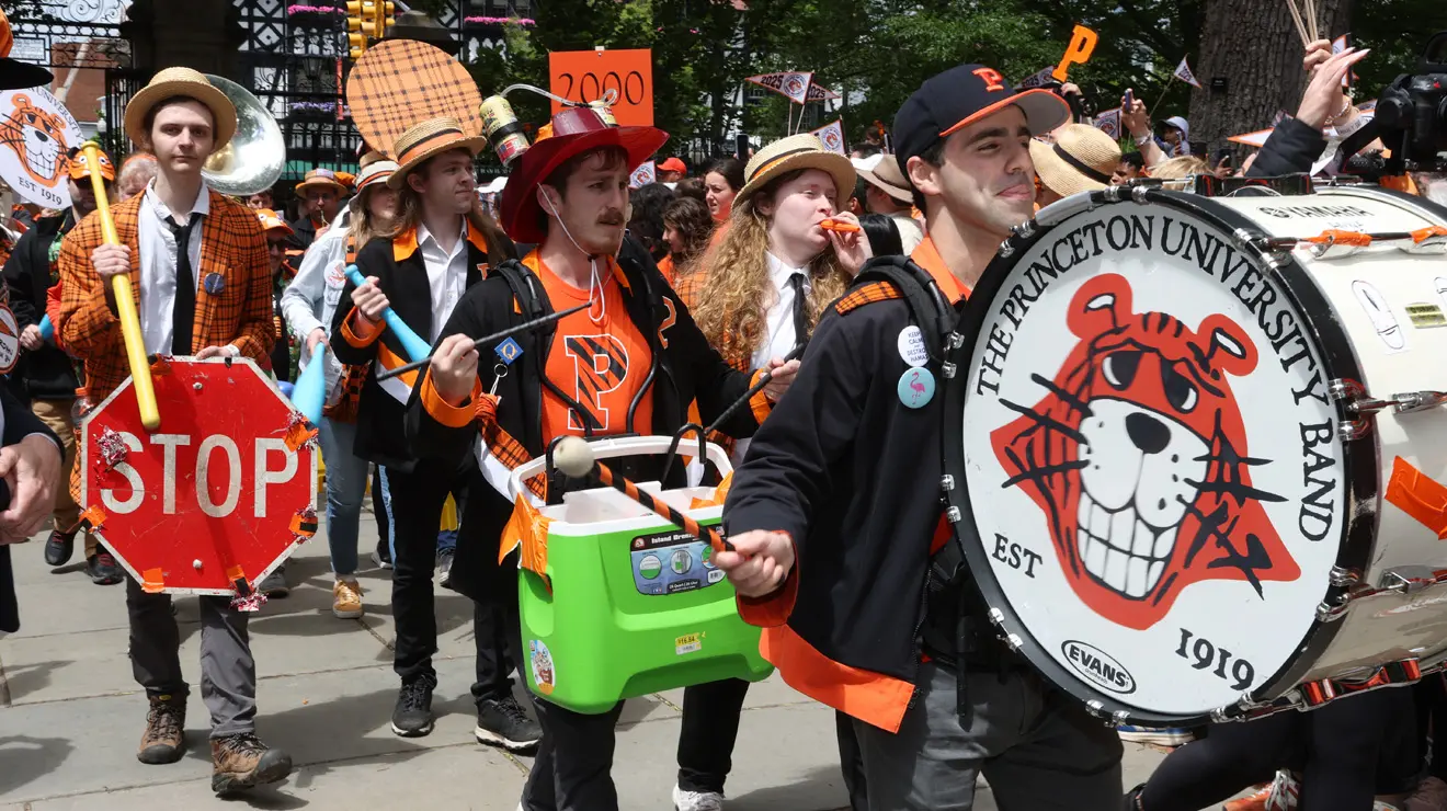 The Princeton University band march in the P-rade; a drummer holding a large drum with a cartoon tiger face on it leads the group.