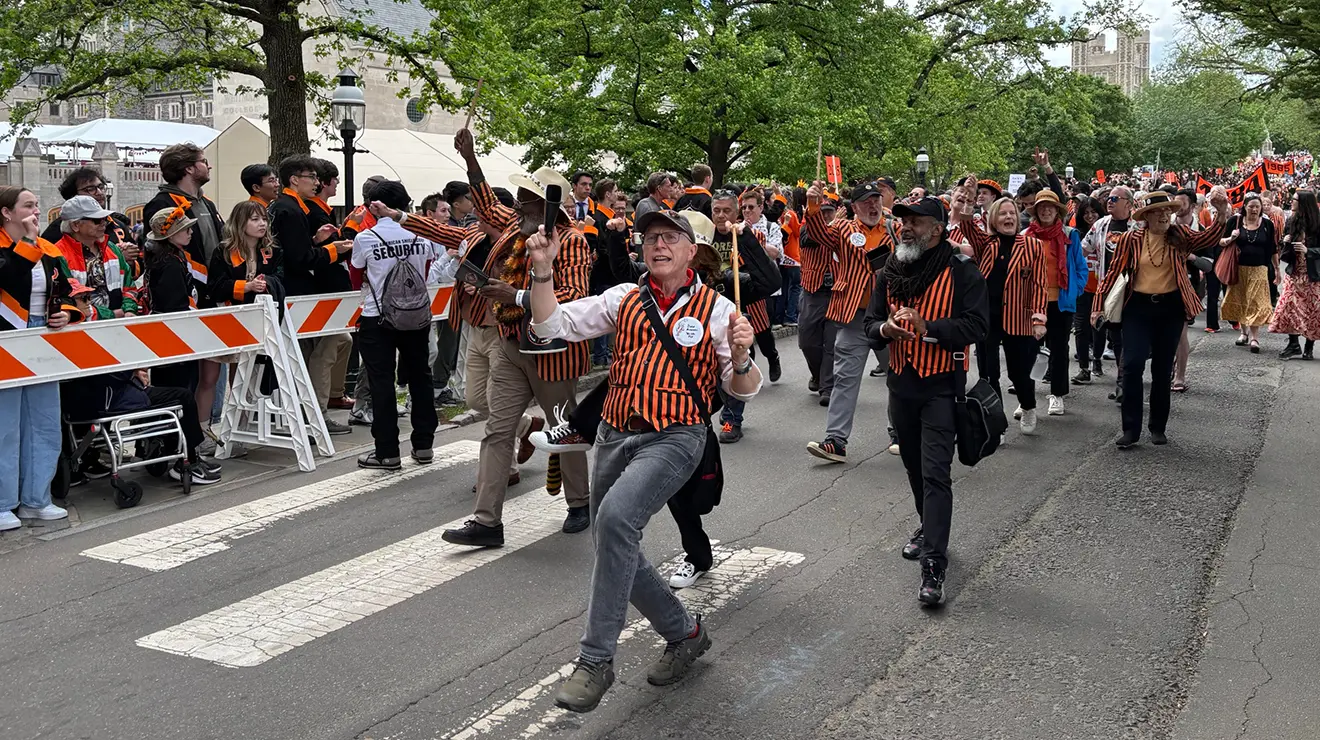 A man in an orange striped vest caught in mid-step as he leads a group of alumni marching in the P-rade.
