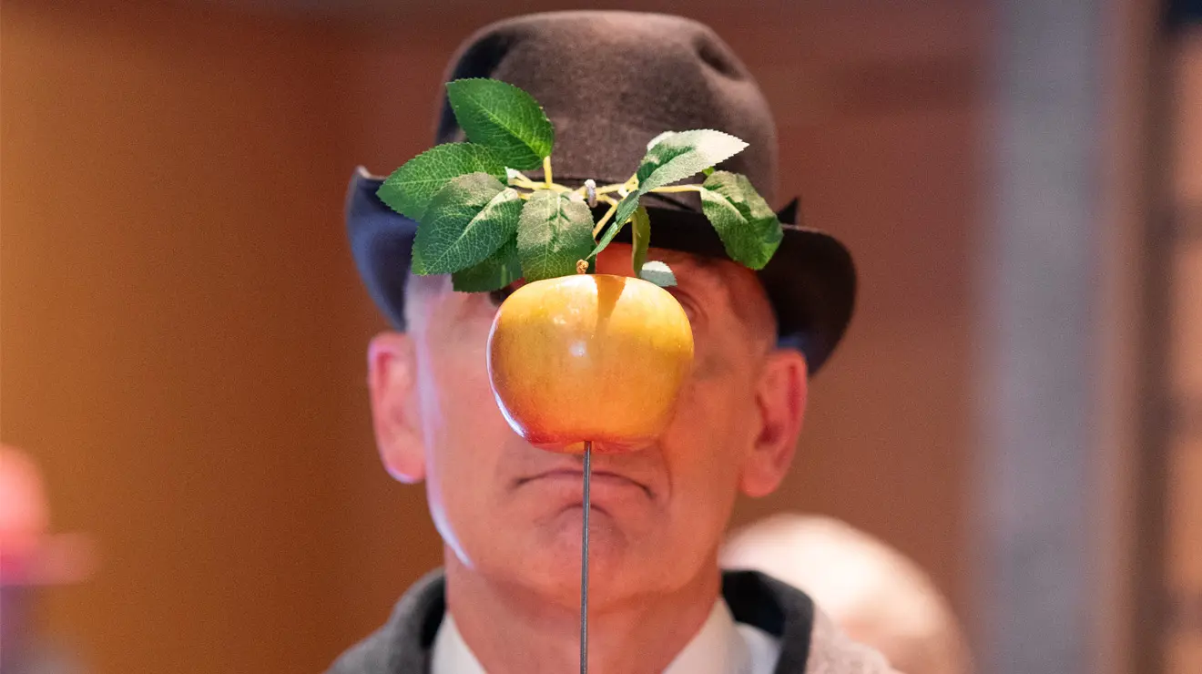PAUM OPENING_Apple.jpg A man wearing a bowler hat holds an apple on a stick in front of his face.