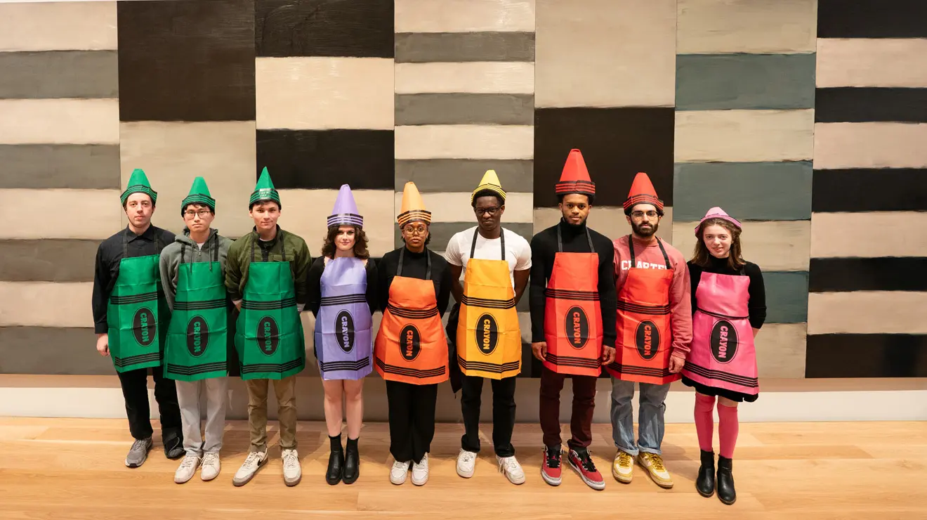 PAUM OPENING_Crayons.jpg Nine people dressed as crayons line up in front of a striped wall.