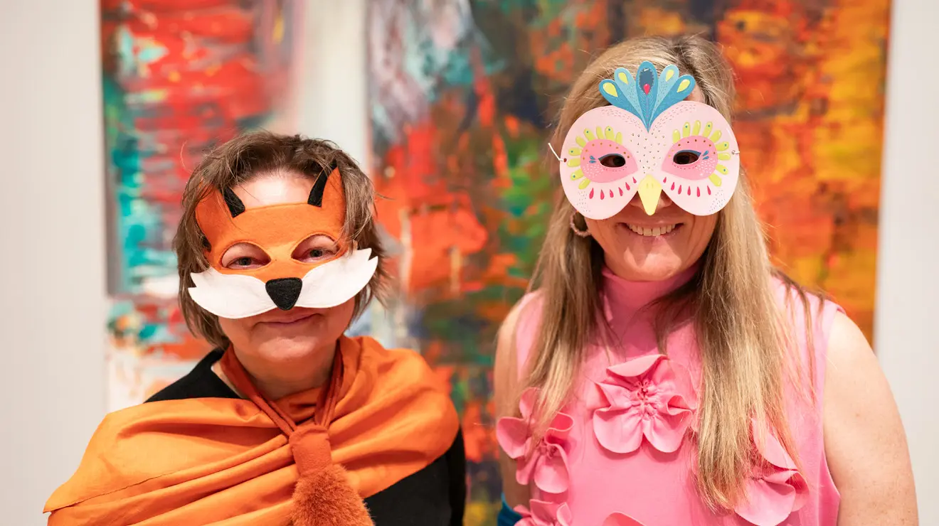 PAUM OPENING_Fox and bird costumes.jpg In front of a colorful painting, two women wear paper face masks on Halloween, one dressed as a fox and the other dressed as a bird.