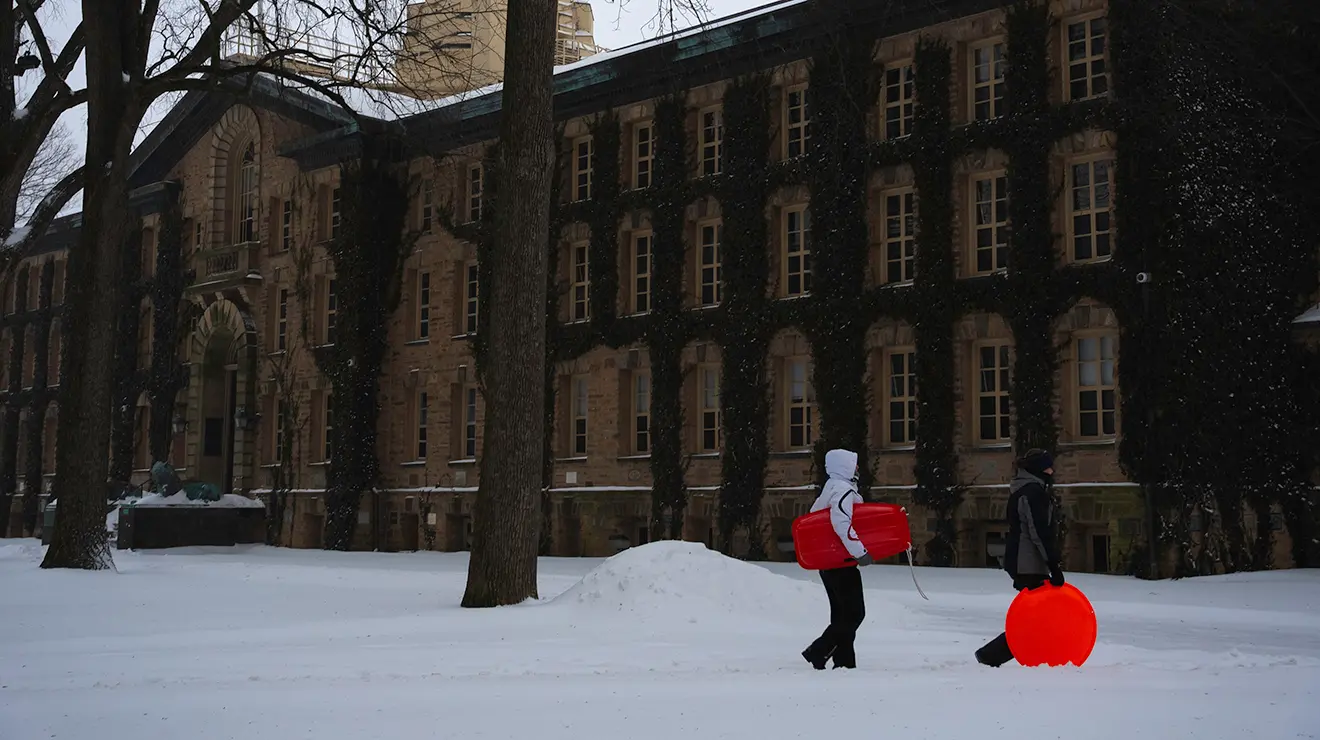 Two people carry red sleds in front of Nassau Hall in the snow.