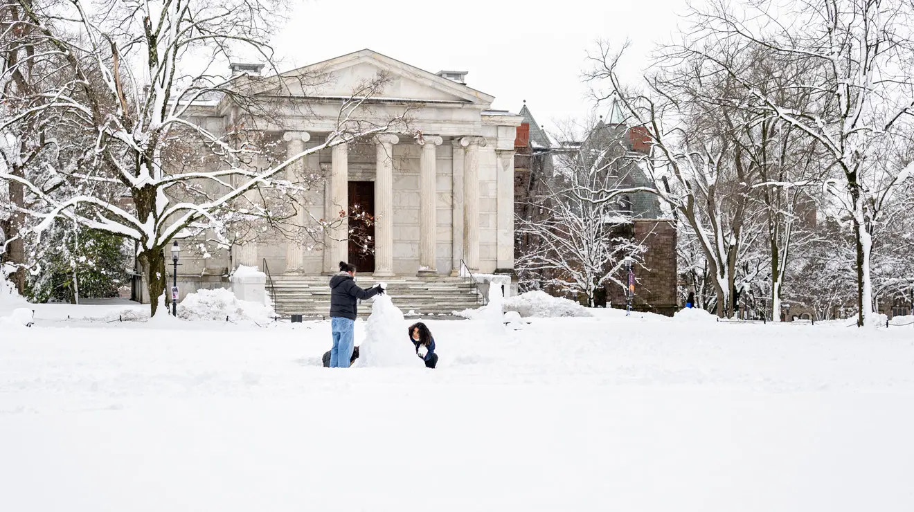 Students build a snowman after a blizzard, with a white columned building in the background.