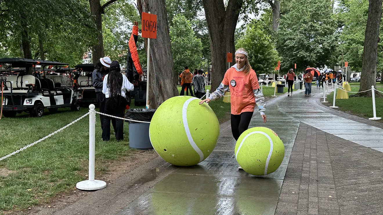 A woman stands between two giant tennis balls, one coming up to her knee and the other above her hip.