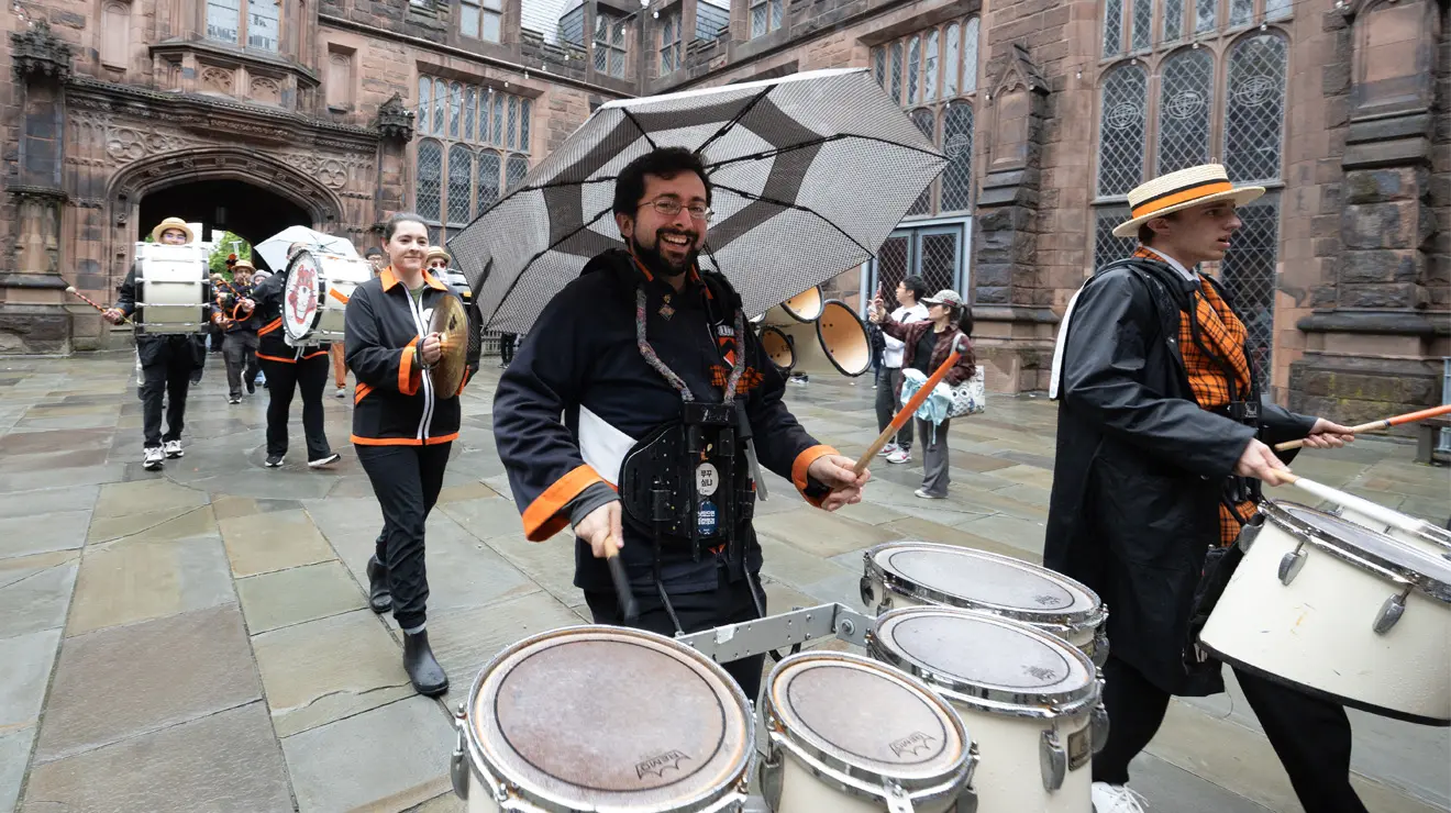 A man plays four drums in a marching band going through the East Pyne building.
