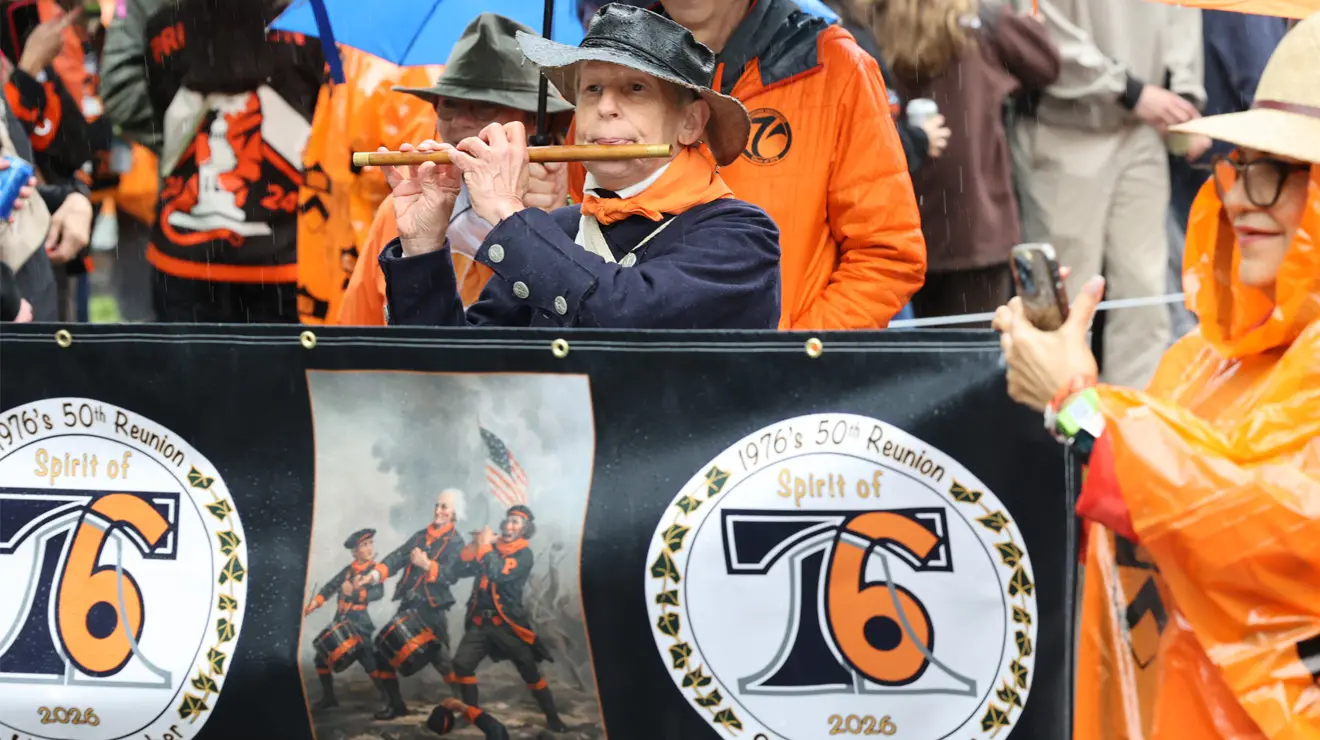 A man plays a fife behind a 1776 banner.