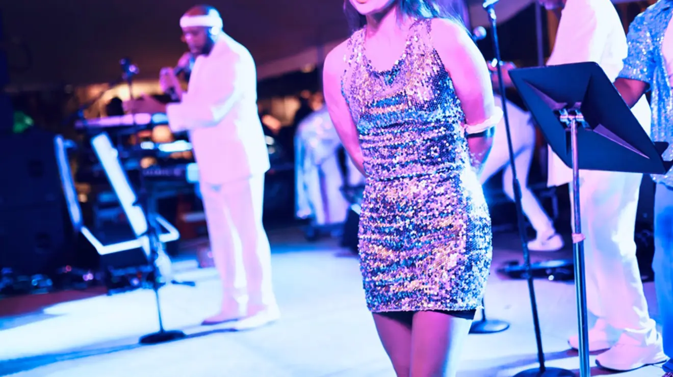A woman on stage in a sparkly dress; men appear to play music in the background.