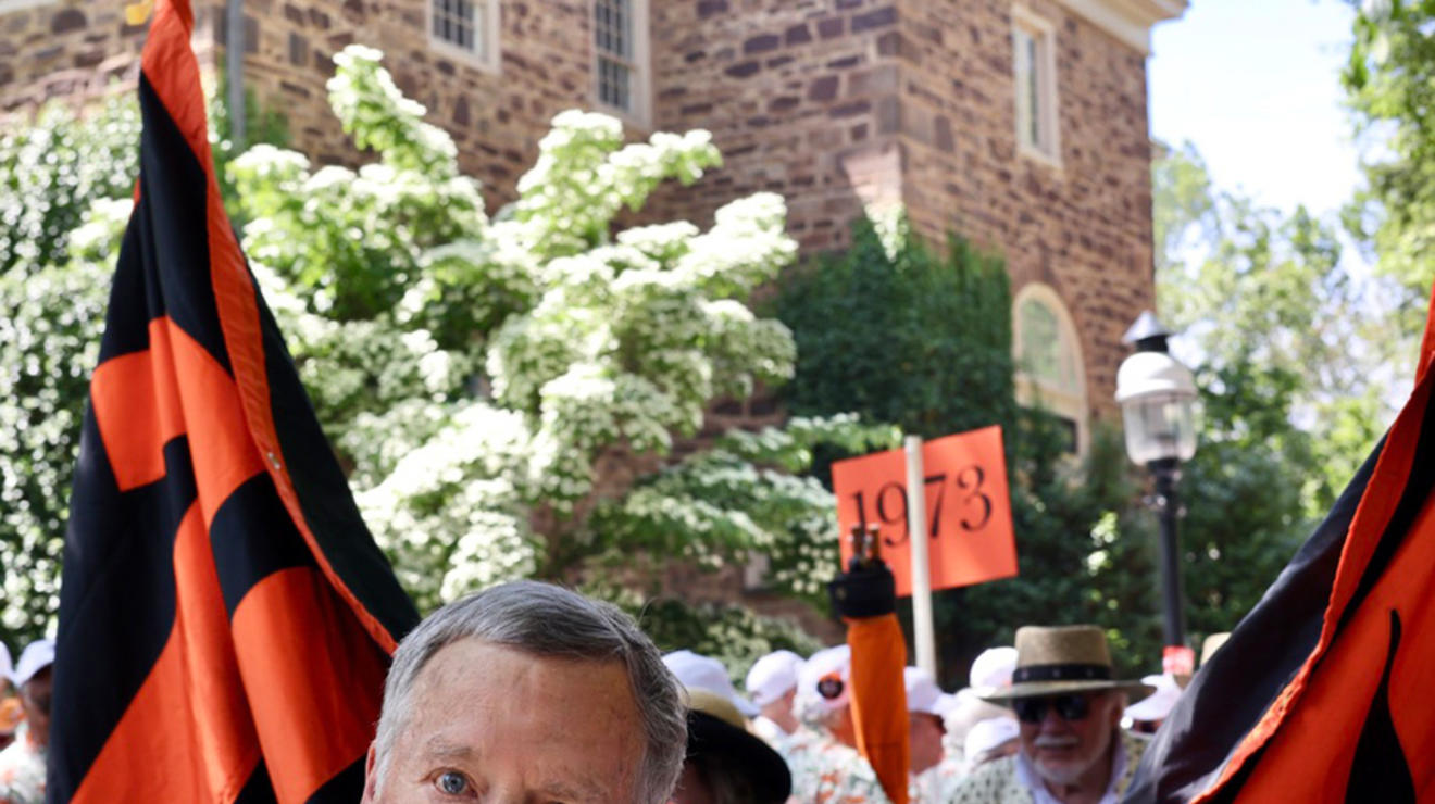 A man peers at the camera near an orange-and-black banner; someone in the background holds a sign reading "1973."