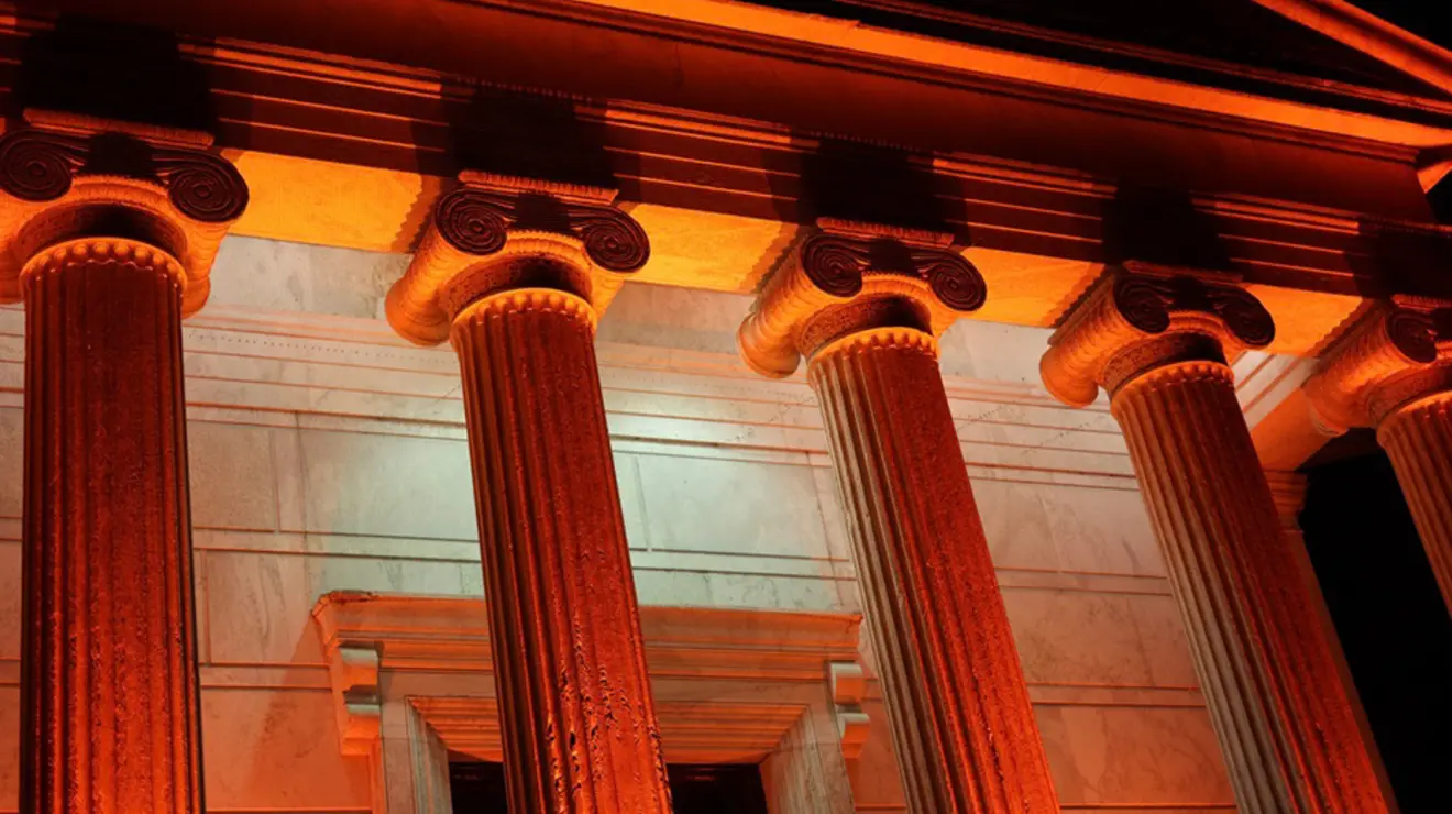 The tops of five Ionic columns on a building are lit up with orange lights.