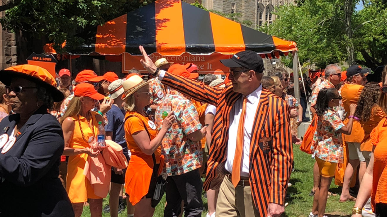 A man wearing a black and orange striped jacket waves to people during the P-rade.