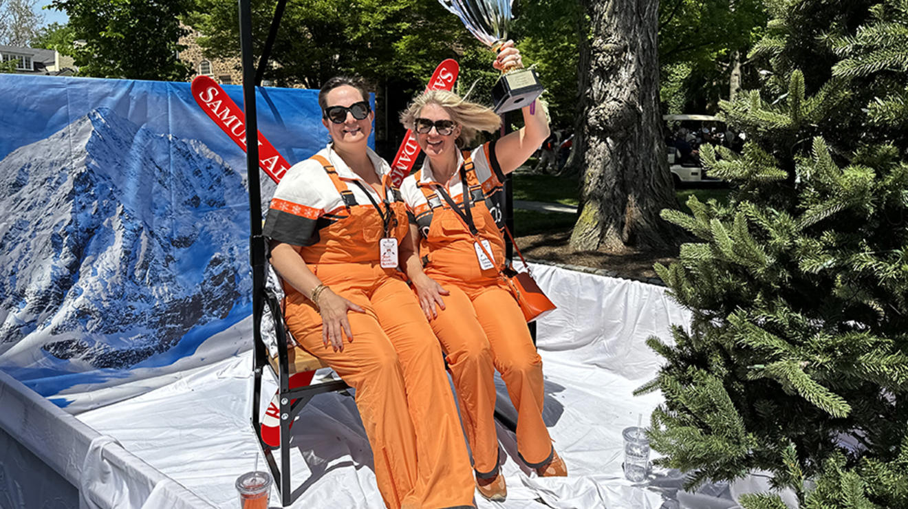 Two women wearing orange overalls; one is holding a trophy over her head.