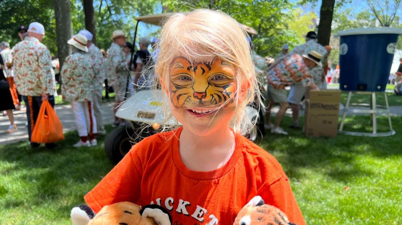 A six-year-old girl with tiger faceprint, an orange shirt, and a plush tiger under each arm.