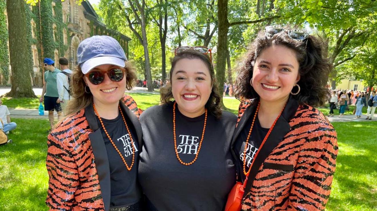 Three women wearing orange martini gras beads and T-shirts reading "The 5th"; the two on the outside are wearing sequined, tiger-striped jackets.