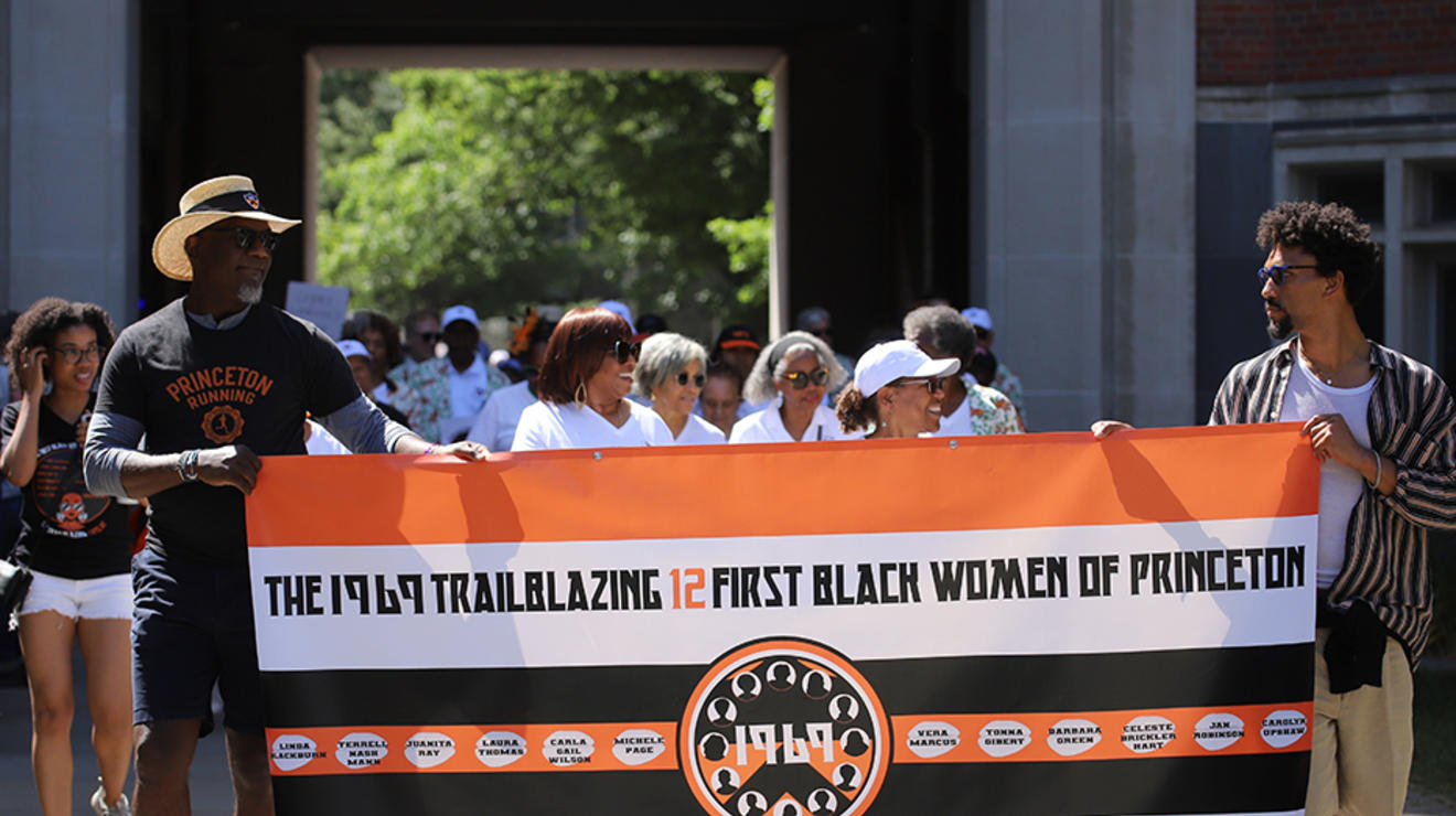 Two guys carry a banner reading "The 1969 Trailblazing 12 First Black Women of Princeton."