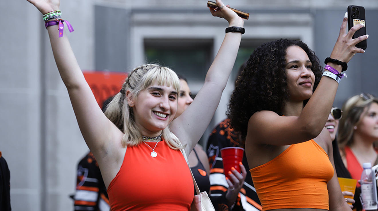 A girl raises up her arms; another girl next to her takes a photo with her cell phone.