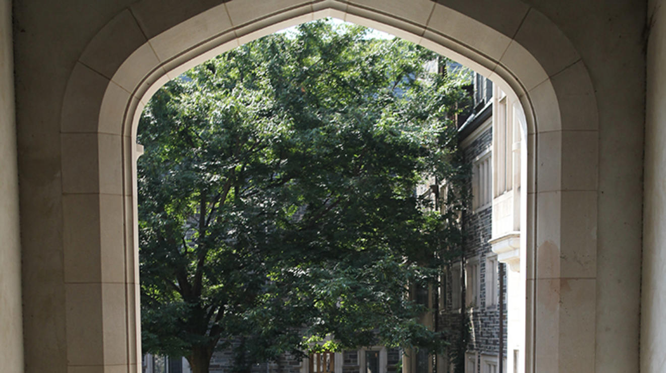 A student pushes a large orange bin on wheels past a stone archway.