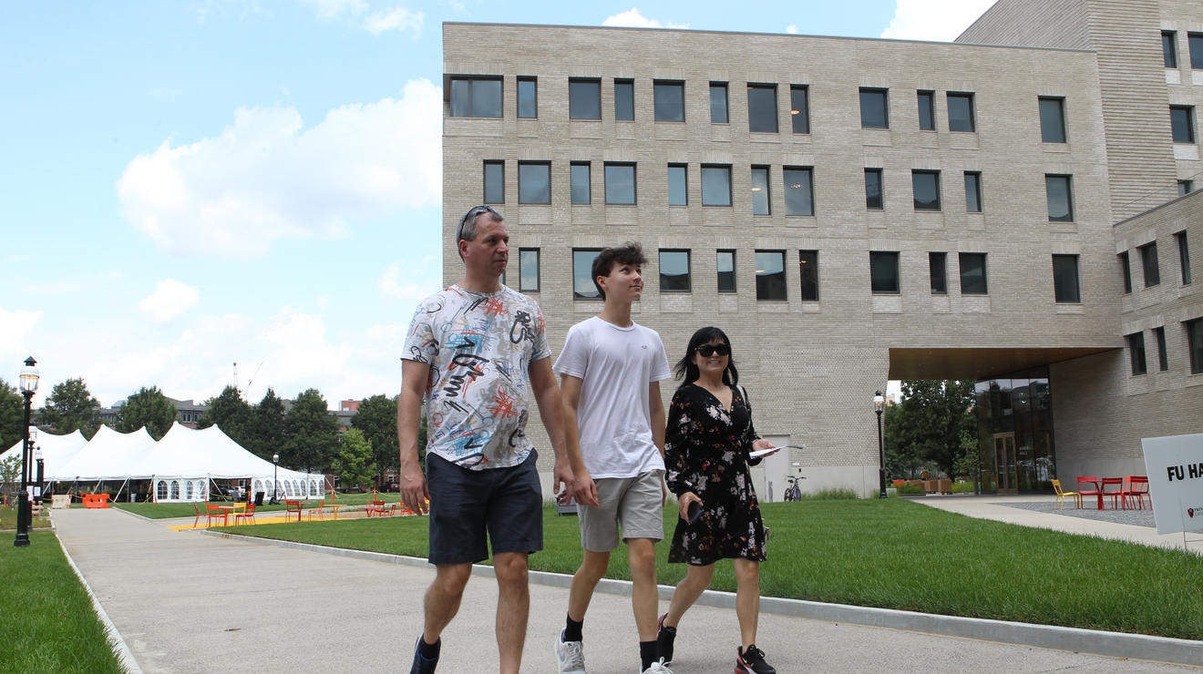 A student and his parents walk down a path with Yeh College and a white tent in the background.